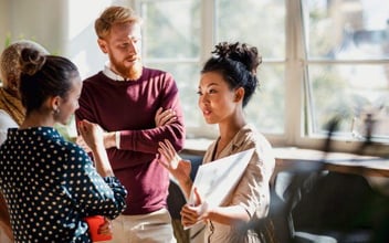 Colleagues standing in a small group discussing something. One of the women is holding documents and gesturing with her hands as the others watch and listen.