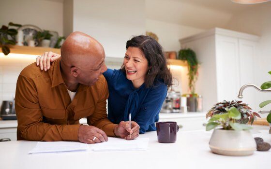 Mature Couple Reviewing And Signing Domestic Finances And Investment Paperwork In Kitchen At Home