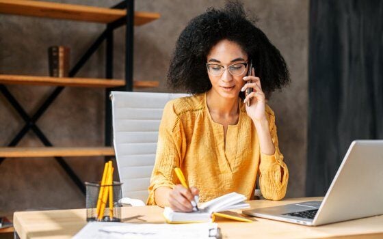 A young a woman is office employee works with a laptop in the office, she takes a notes while talking by phone