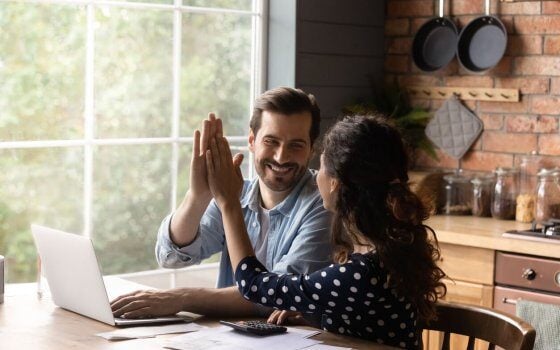 Happy young family couple giving high five to each other, finishing managing household budget, celebrating successful investment, making all payments online in e-banking computer application.