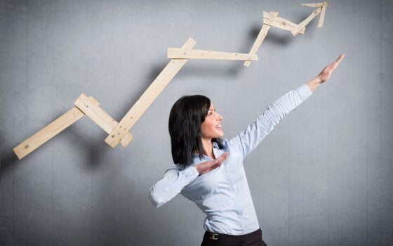 Concept: Successful business trend. Happy talented businesswoman pointing arm upwards in front of ascending business graph, isolated on grey background.