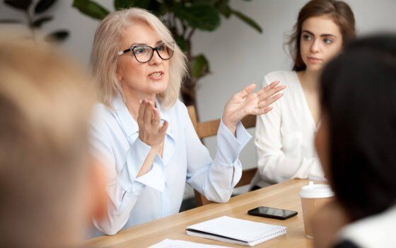 Attractive aged businesswoman, teacher or mentor coach speaking to young people, senior woman in glasses teaching audience at training seminar, female business leader speaker talking at meeting