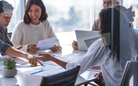 Multi racial diverse group of people working with Paperwork on a board room table at a business brainstorm,  presentation or seminar. The documents have financial or marketing figures, graphs and charts on them. People are pointing to different documents. There are laptops and digital tablets on the table. They are happy and smiling.