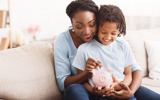 Money Saving Concept. Excited black mom and daughter putting coins into piggy bank, free space