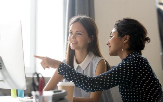 Smiling millennial diverse colleagues discussing online work together, happy caucasian female intern listening to indian mentor explaining computer task training employee, office mentoring concept