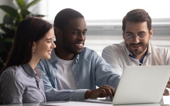 Smiling african american team leader sitting at table with colleagues, working on computer at workplace. Diverse young teammates looking at positive economic growth results report at office.