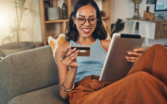 Smiling mixed race woman using credit card for ecommerce on digital tablet at home. Happy hispanic sitting alone on living room sofa, using technology for ebanking. Relaxing, ordering, buying online