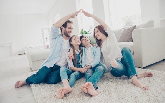 New building residential house purchase apartment concept. Stylish full family with two kids sitting on carpet, mom and dad making roof figure with hands arms over heads