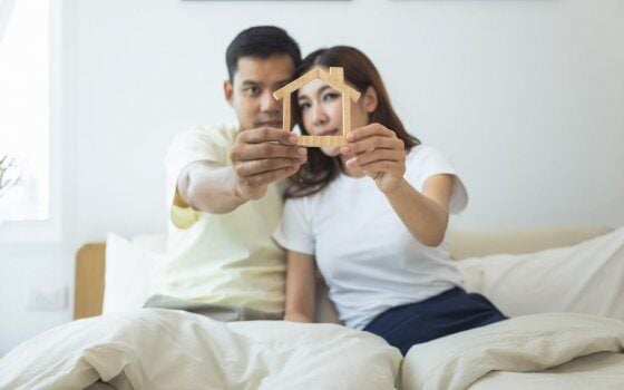 Couple or 2 asian people on bed in bedroom, home or house. Man and woman hold home model in hand together. Look happy, romantic. Concept for married, family, rent, buy, real estate and propertry.