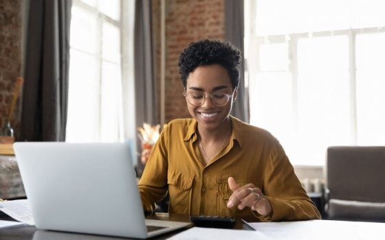 Happy young Afro American entrepreneur woman in glasses counting profit, on calculator at laptop computer, analyzing benefits, enjoying financial success, job high result, smiling