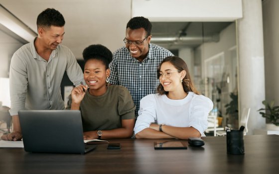 Diverse employees gathered in office having fun watching funny video, discussing new ideas, brainstorming using laptop enjoying break in workday