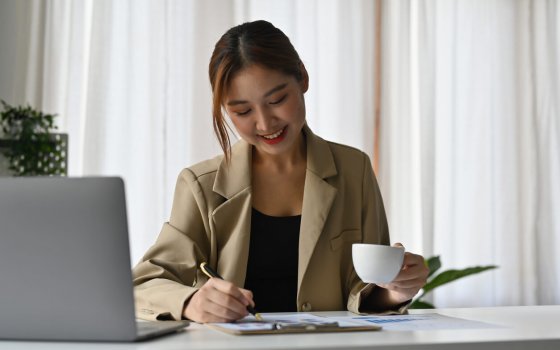 Smiling young cheerful Asian woman  enjoying morning coffee while working on the laptop at modern office.