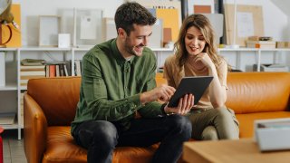 Smiling business man and business woman sitting on a orange sofa and working together on a digital tablet