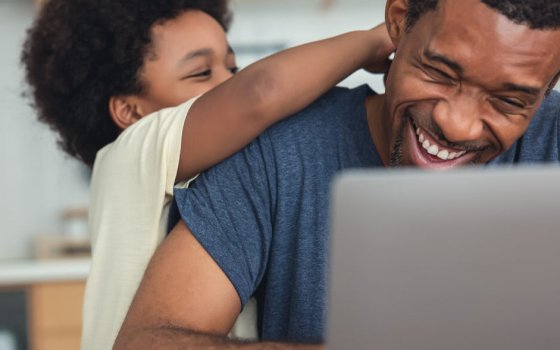 Cute little African American boy hugging and playing with dad while his dad working from home with laptop computer for remoter job in modern kitchen. Concepts of remote work, children, parenthood